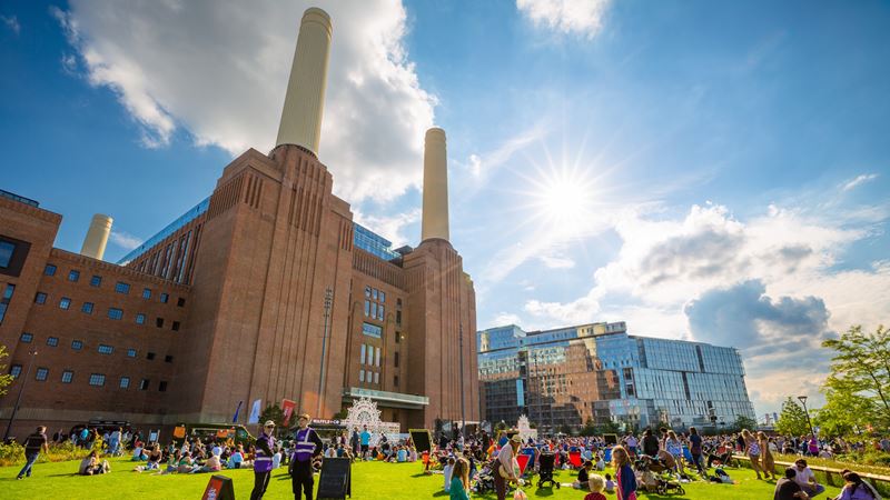 Battersea Power Station chimneys with people sitting on the grass in front and blue sky with sunshine.
