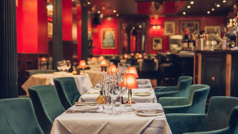 Enjoy an elegant setting to enjoy classic British fare at Boisdale of Belgravia. Credit: Boisdale of Belgravia. Image courtesy of Boisdale of Belgravia. Dining tables lined up with white tablecloth, wine glasses, green velvet seating, cosy lighting and artwork on red walls.