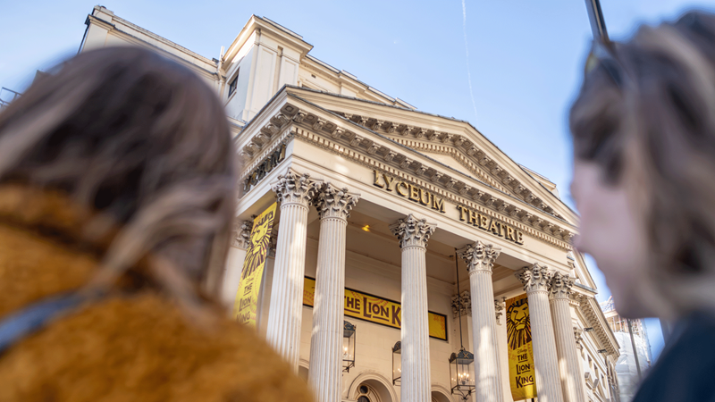 Two women look up at the Lyceum Theatre lined by Lion King advertisements