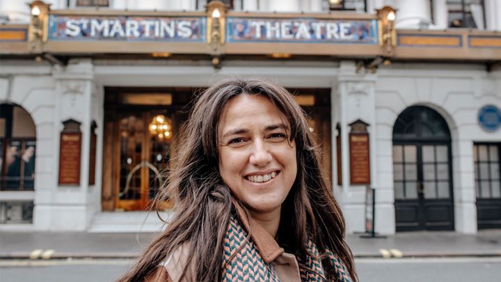 landscape photo of a long, dark brown haired lady wearing a brown coat and scarf, standing smiling in front of St Martins Theatre