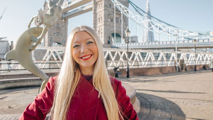 landscape photo of a long, blonde haired lady wearing a red coat, standing smiling in front of Tower Bridge