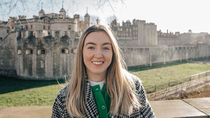 landscape photo of a blonde haired lady wearing a green cardigan buttoned and a chequered coat over the top, standing smiling in front of the Tower of London