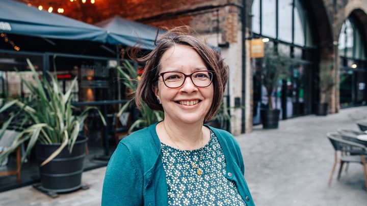 landscape photo of a short brown haired lady wearing a green patterned dress and cardigan, standing smiling in Borough Yards