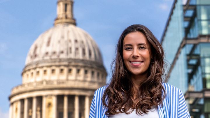 landscape photo of a long, dark brown haired lady wearing a white top and stripey blue overshirt, standing smiling in front of St Paul's Cathedral