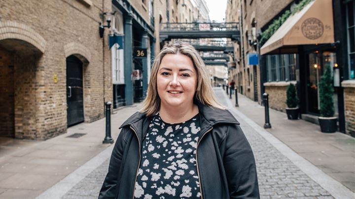 landscape photo of a short blonde haired lady wearing a black and white patterned dress and black coat over the top, standing smiling in the historic riverside street Shad Thames