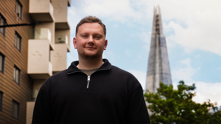 landscape photo of a brown haired man wearing a black half zip, standing smiling in the street with The Shard in the background
