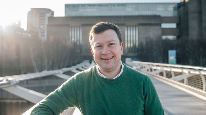 landscape photo of a short brown haired man wearing a dark green jumper, standing smiling on the Millennium Bridge