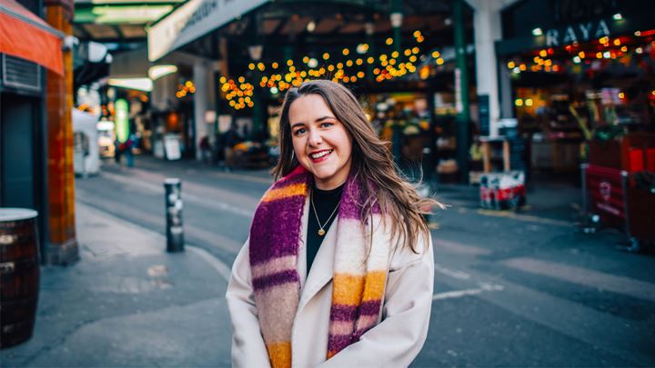 landscape photo of a long, dark brown haired lady wearing a white coat and stripey purple, orange and white scarf, standing smiling in front of Borough Market