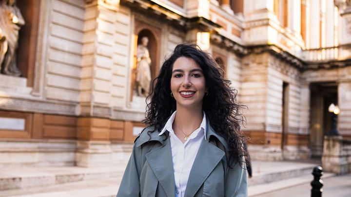 Landscape photo of a long, dark haired lady wearing a white shirt and khaki green trench coat, standing smiling in front of the Royal Academy of Arts.