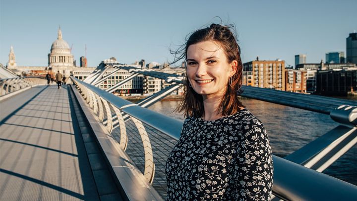 landscape photo of a short brown haired lady wearing a black and white spotty dress, standing smiling on the Millennium Bridge