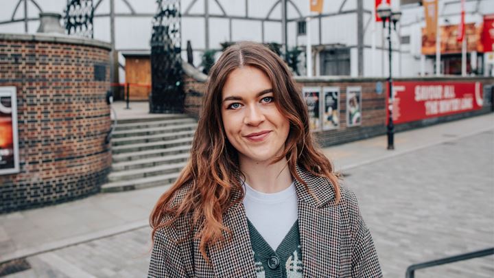 landscape photo of a long, dark brown haired lady wearing a green cardigan and Herringbone-print coat over the top, standing smiling in front of Shakespeare's Globe