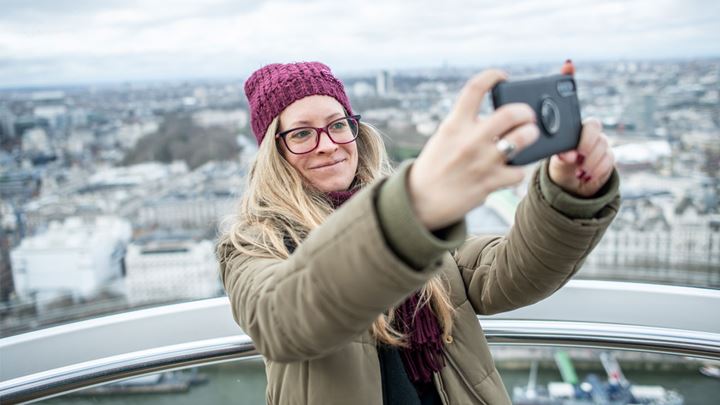 landscape photo of a long, blonde haired lady wearing a green coat and purple hat, holding a phone up to take a selfie on the London Eye
