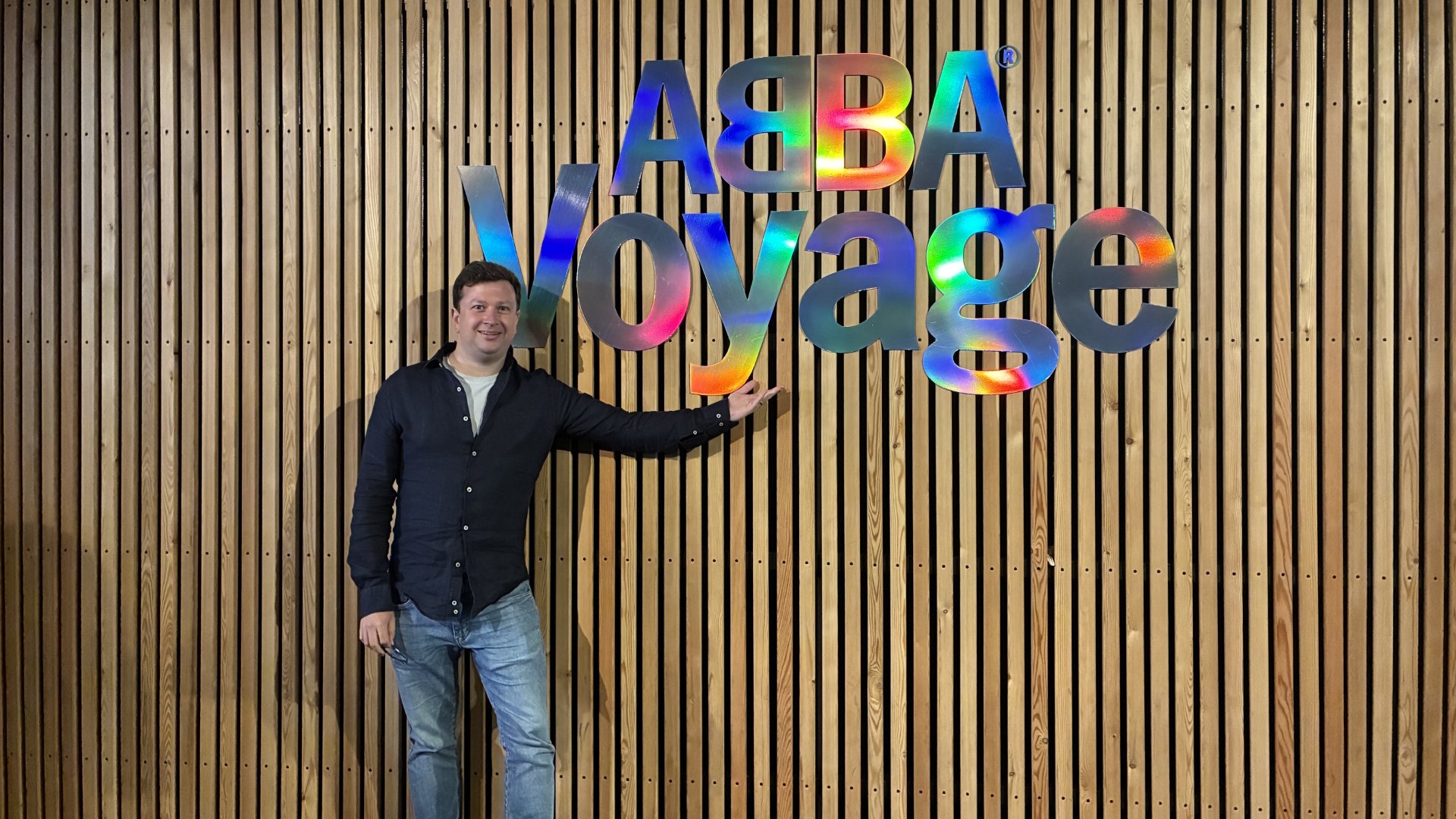 Take a picture with the ABBA Voyage logo inside the ABBA Arena concourse before the show. Image courtesy of Jonny Payne. A man stands infront of a wooden wall displaying the multicoloured, shiny, ABBA Voyage logo.