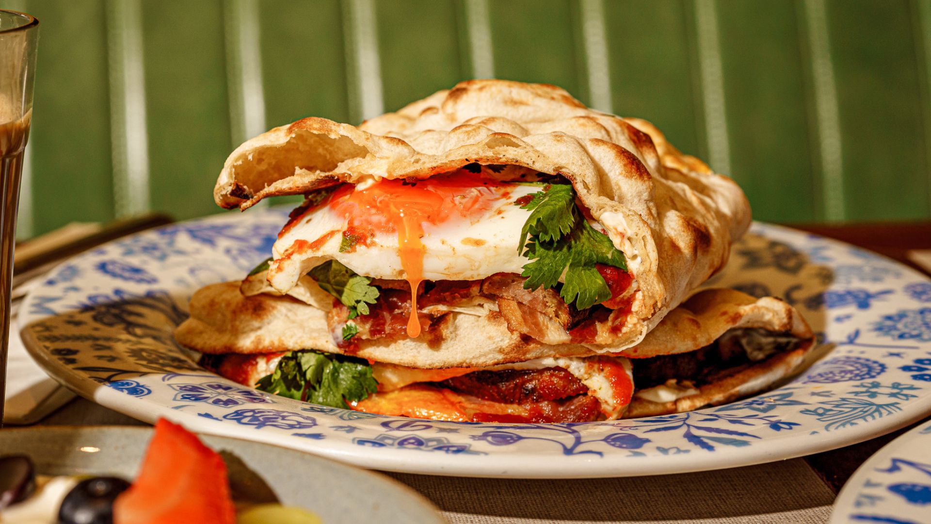 Try Dishoom's Wrestler Naan, freshly baked naan with smoked bacon, pork sausages and a runny-yolked egg. Image courtesy of Dishoom. a naan bread stuffed with bacon, sausage and a runny yolked egg, presented on a small flowery white and blue plate