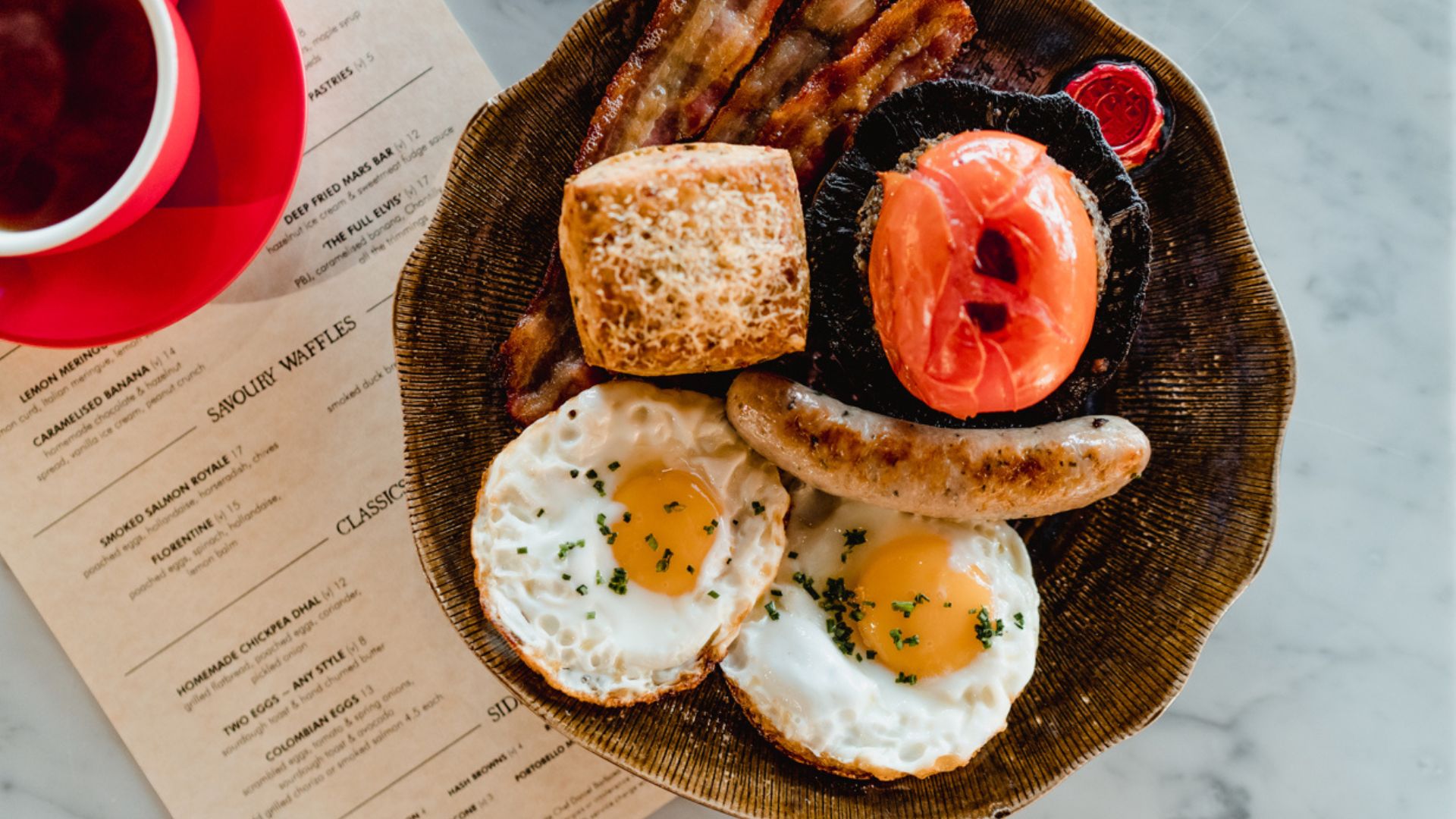 Treat yourself to a Duck & Waffle English breakfast and panoramic views across London from the 48th floor. Image courtesy of Duck & Waffle. high angle photo of a small plated English breakfast with sausage, eggs, bacon, tomato, cheese scone and a cup of tea just out of shot