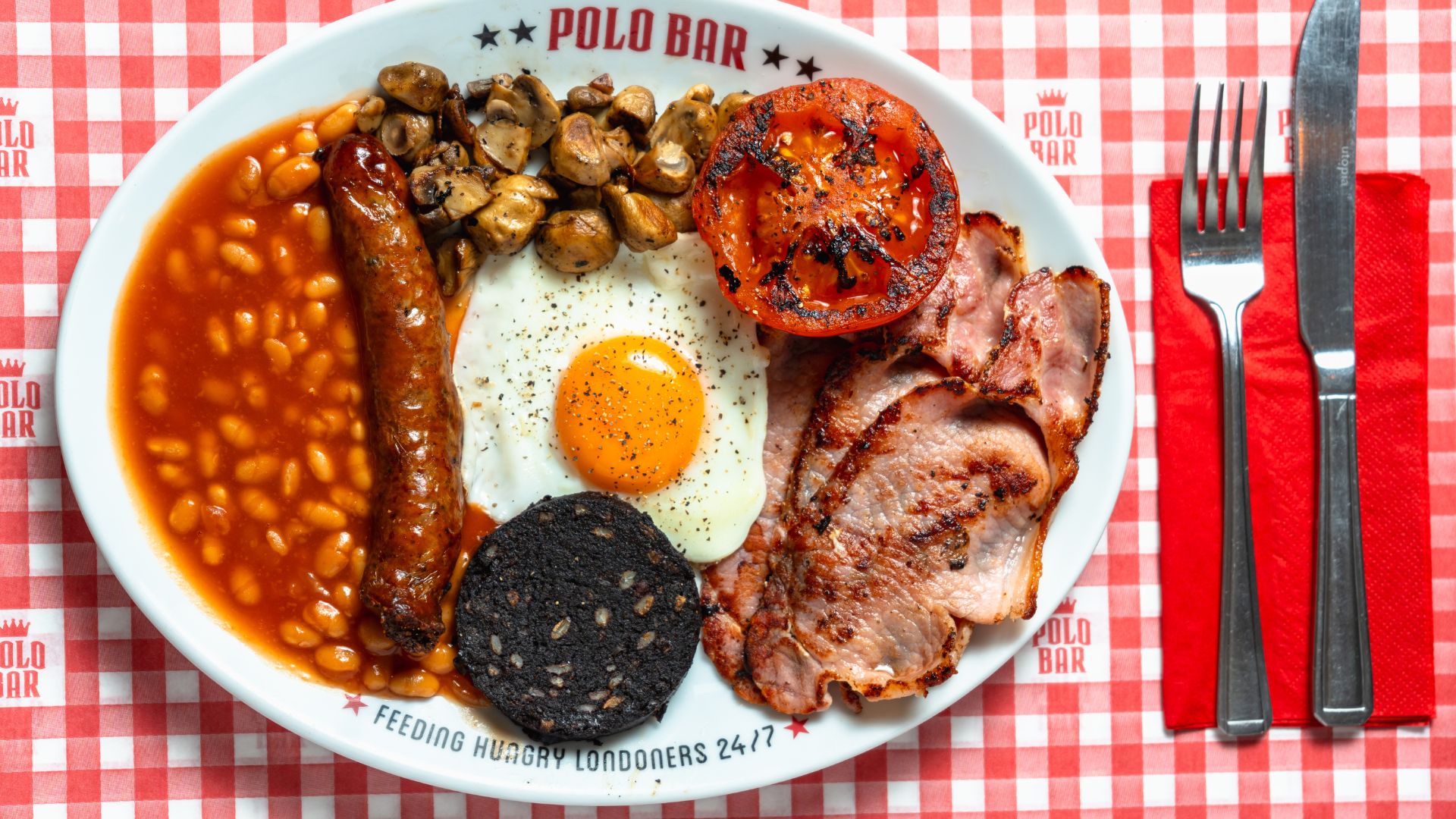 Visit Polo Bar for English breakfasts served 24 hours a day. Image courtesy of Polo Bar. high angle photo of a white plate of beans, sausage, egg, bacon, mushrooms, tomatoes and black pudding, on a chequered red and white tablecloth