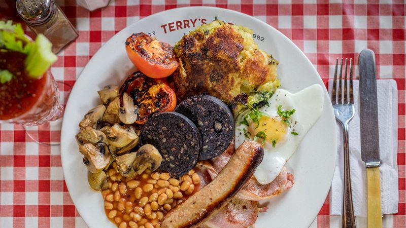 high angle photo of a white plate of beans, sausage, egg, bacon, mushrooms, tomatoes, bubble and squeak and black pudding, on a chequered red and white tablecloth