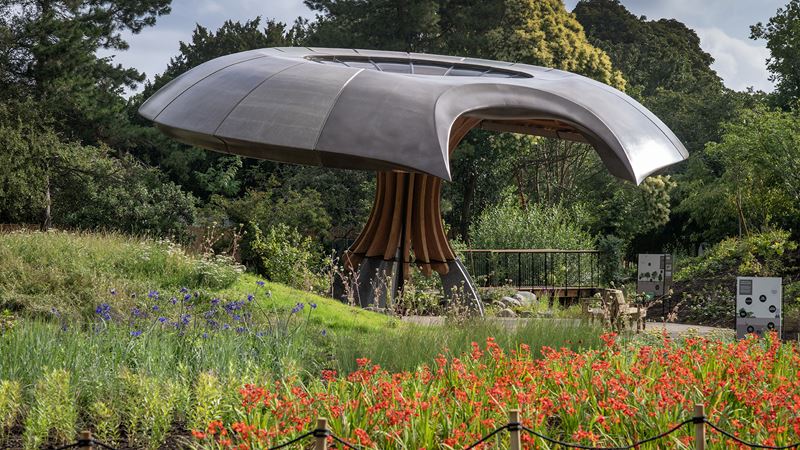 An image of a mushroom like pavilion in the center of the Carbon Garden at Kew surrounded by green grass, trees and red flowers.