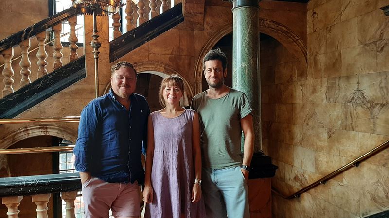 Three members of the Harry Potter and the Cursed Child cast stand at the staircase next to one of the green columns within the Palace Theatre.