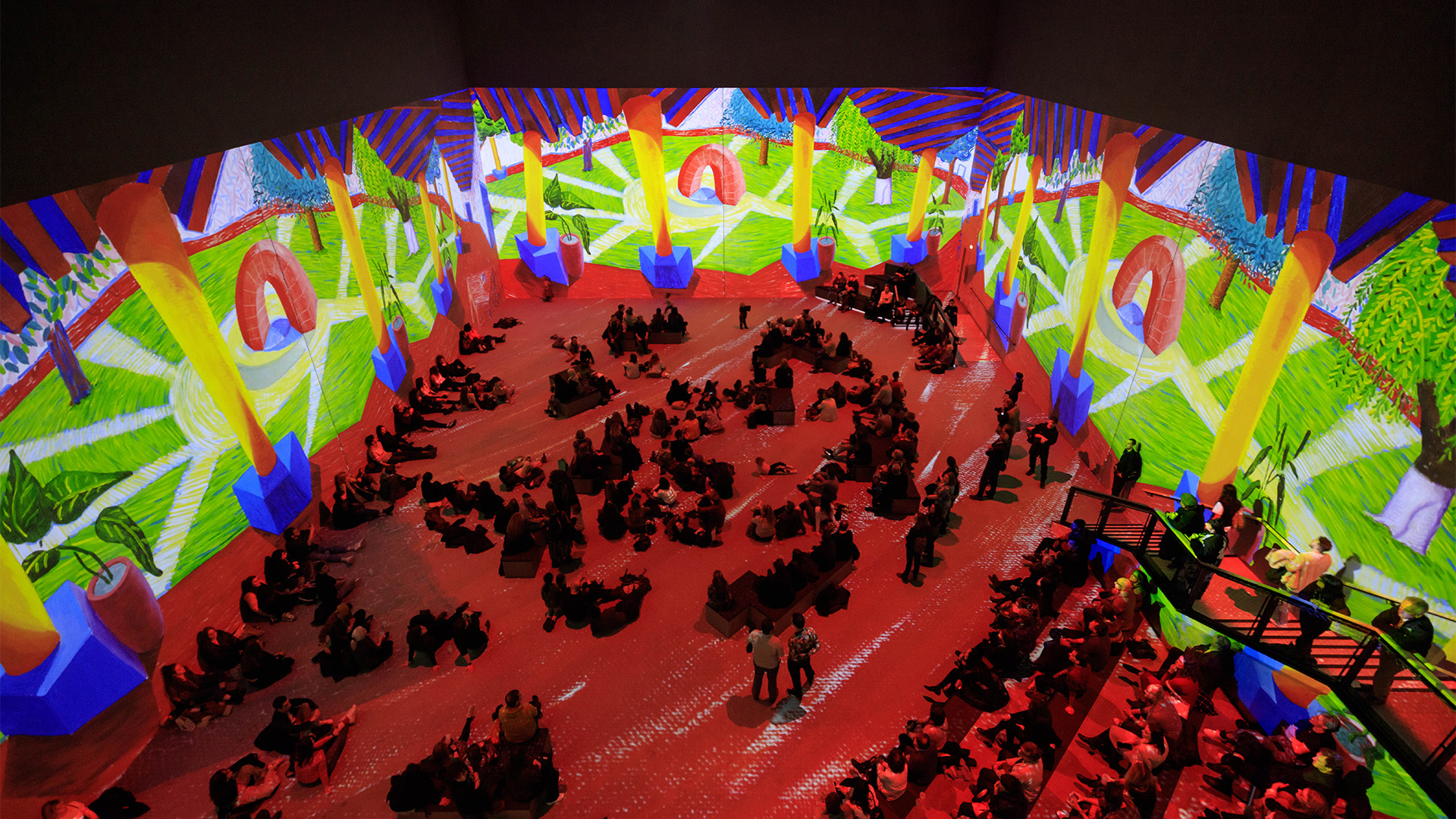 Audiences engage with wraparound Hockney artworks at his Lightroom exhibition. Image courtesy of Lightroom. Wraparound screens project a bright, bold landscape painting by David Hockney at Lightroom.