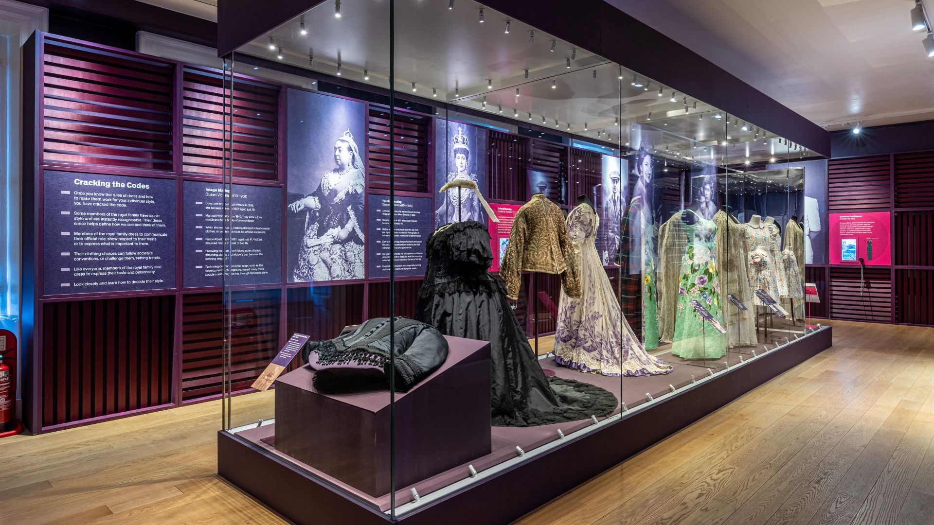 Discover garments worn by Queen Elizabeth II, Queen Victoria, Princess Diana and other fashionable royals at Kensington Palace. © Historic Royal Palaces. Glass cabinet showing royal outfits with pictures and descriptions on the wall behind.