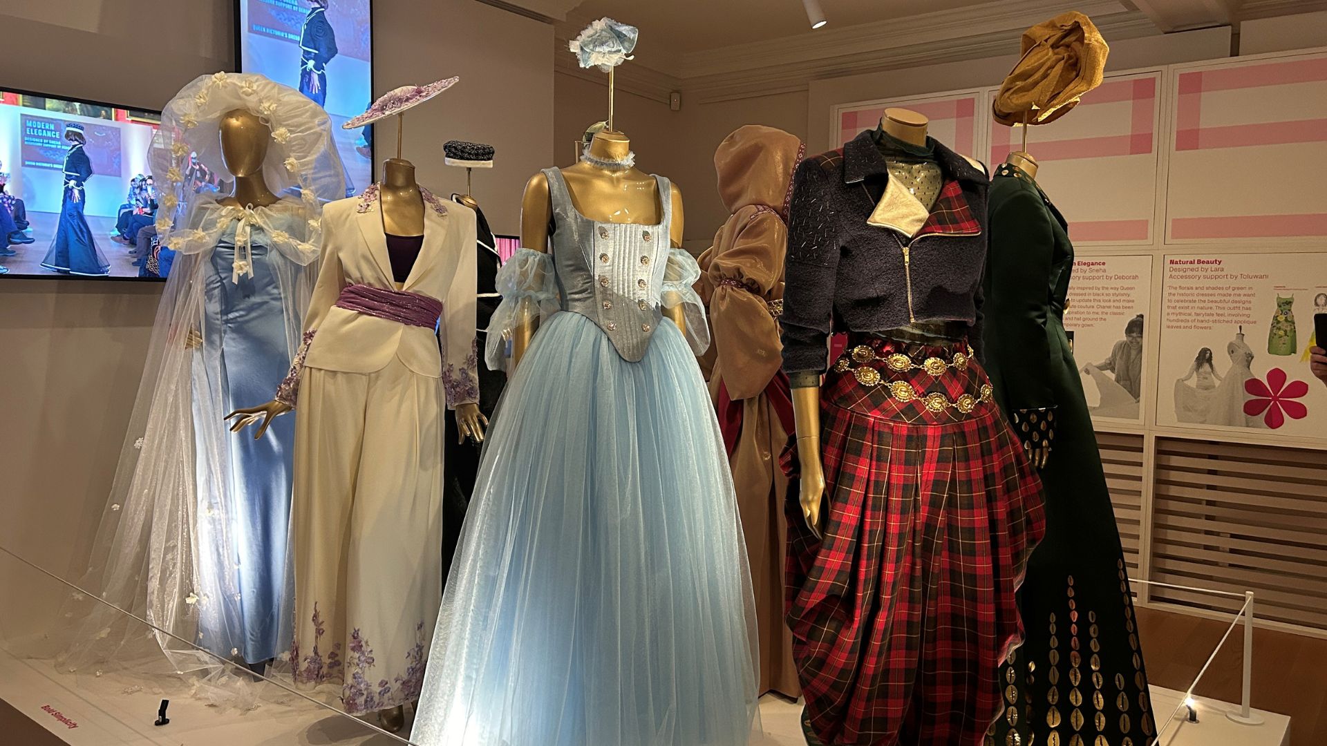 Discover contemporary garments at Kensington Palace designed by the Young Producers. © Visit London/Charlotte Pritchard. Multiple garments lined up in an exhibition room with a tartan and blue dress at the front.