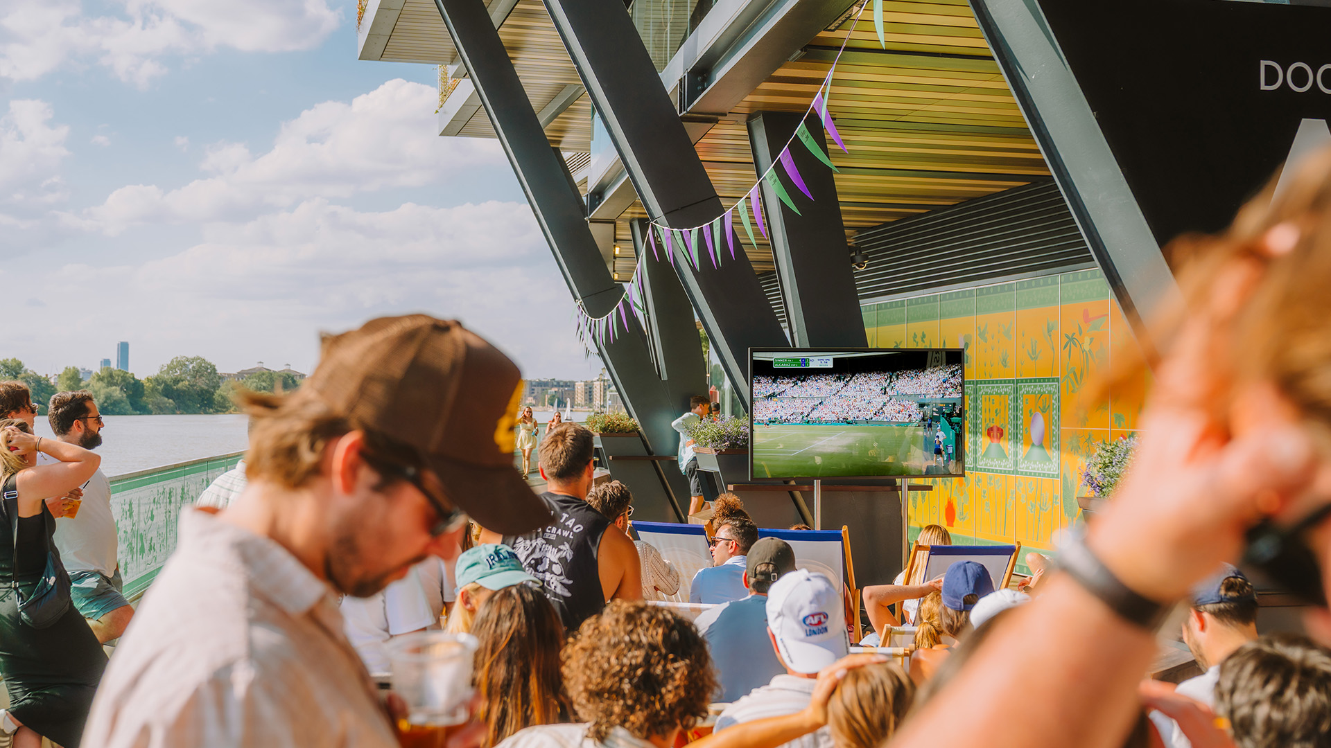 Crowds enjoy a screening of Wimbledon on the sun-drenched boardwalk of Fulham Pier. Image courtesy of Fulham Pier. People watch Wimbledon on a TV screen at Fulham Pier