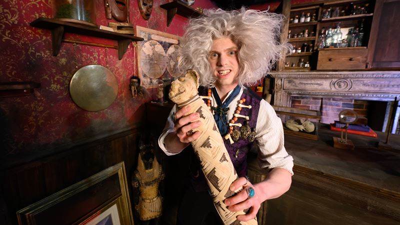 Man with grey wig and Victorian-period clothing holding a mummified object at the London Dungeon in dark-lit room.