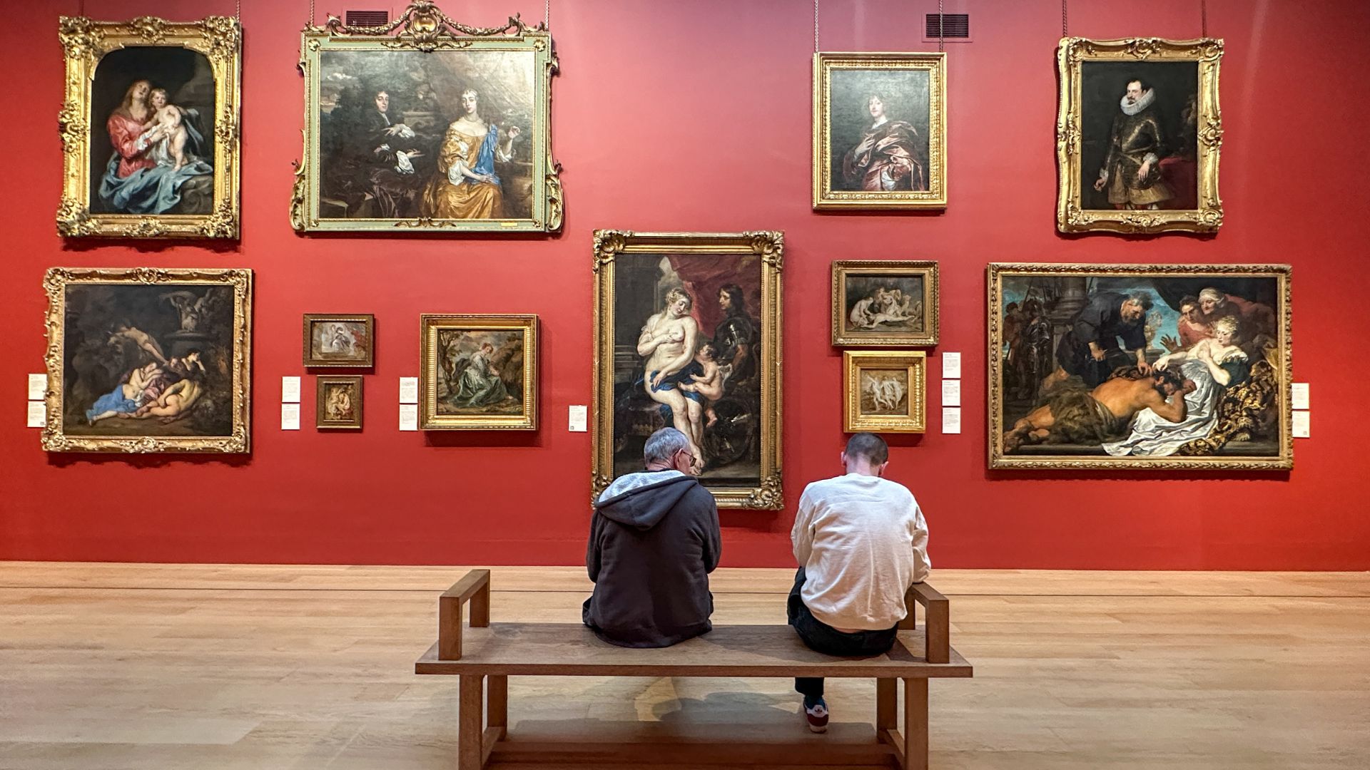 Enjoy world-class art at Dulwich Picture Gallery in south London. Image courtesy of Kirstine Spicer. Two men sit on a brown bench looking at a red wall covered in paintings.