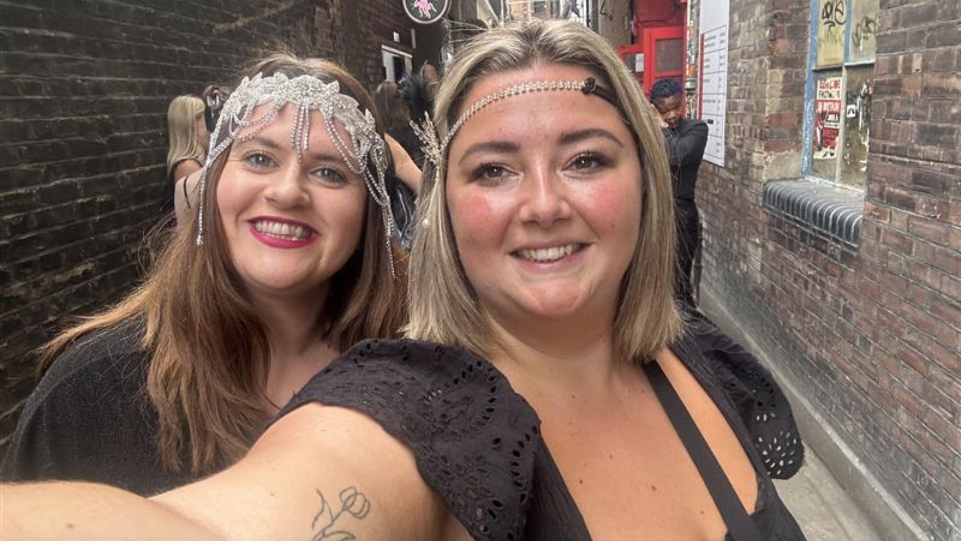 Step into London’s secret speakeasy in 1930s New York at 58th Street. Photo credit: Hannah Cornish two women holding up a phone smiling and taking a selfie; both are wearing black dresses and wearing a 1930's-style feather and gem headband