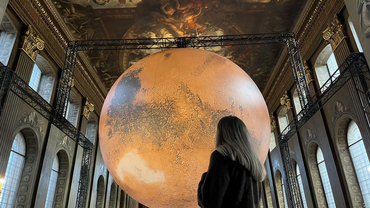Experience Luke Jerram's Mars installation at the Painted Hall, Old Royal Naval College. Image courtesy of Charlotte Pritchard. A lady looks up at the large Mars installation, a huge red sphere that looks like the planet, within the Painted Hall in the Old Royal Naval College.