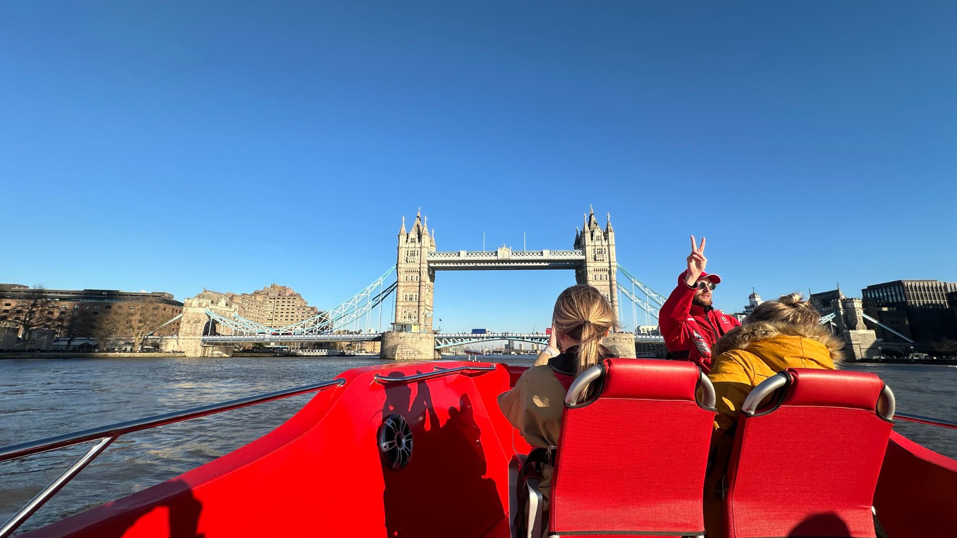 Whizz along the river on a Thames Rockets Speedboat tour. Image credit: Kirstine Spicer. A man wear a red life jacket makes a peace sign in front of tower bridge in london on a red speedboat