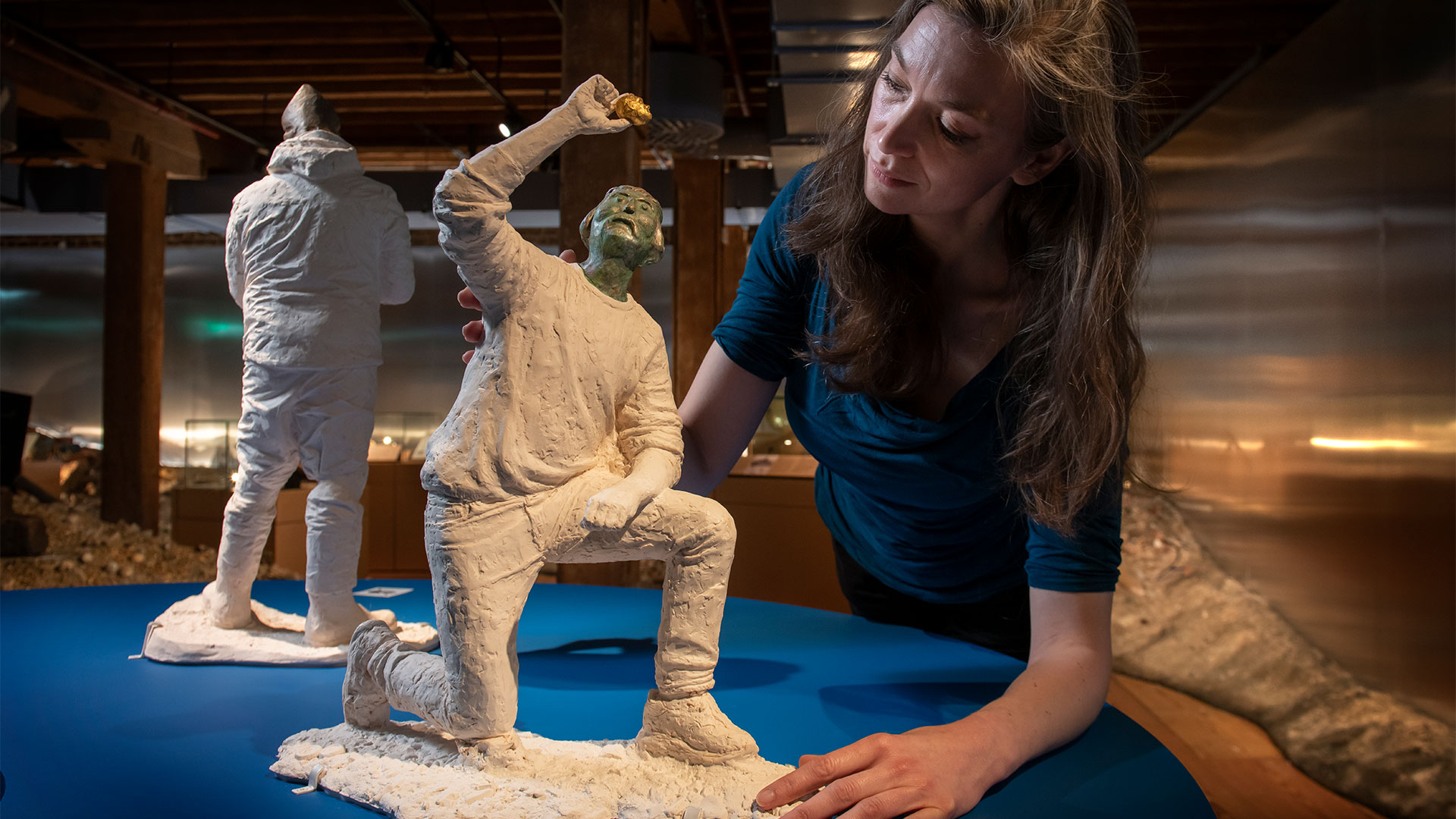 London Museum curator Kate Sumnall pictured with Finders Keepers by sculptor Billie Bond at London Museum Docklands’ Secrets of the Thames exhibition. © London Museum Curator Kate Sumnall stands beside a stone sculpture of a mudlark gazing intently at a golden object discovered in the River Thames.