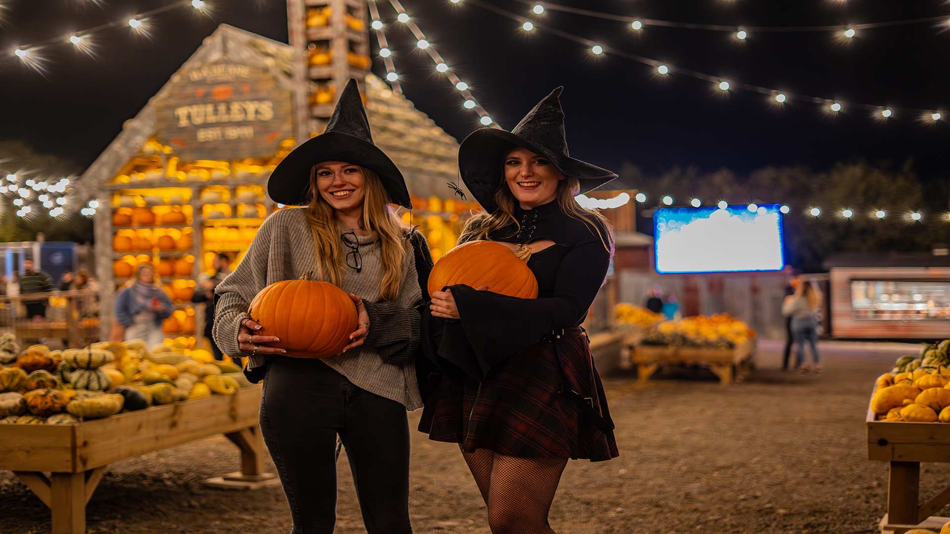 Spend a spooky Halloween at Tulleys Farm and pick your own pumpkin. Image courtesy of Tulleys Farm. Two women dressed with a witch hat holding a pumpkin at nighttime with a farmhouse in the background and lots of stands with pumpkins.
