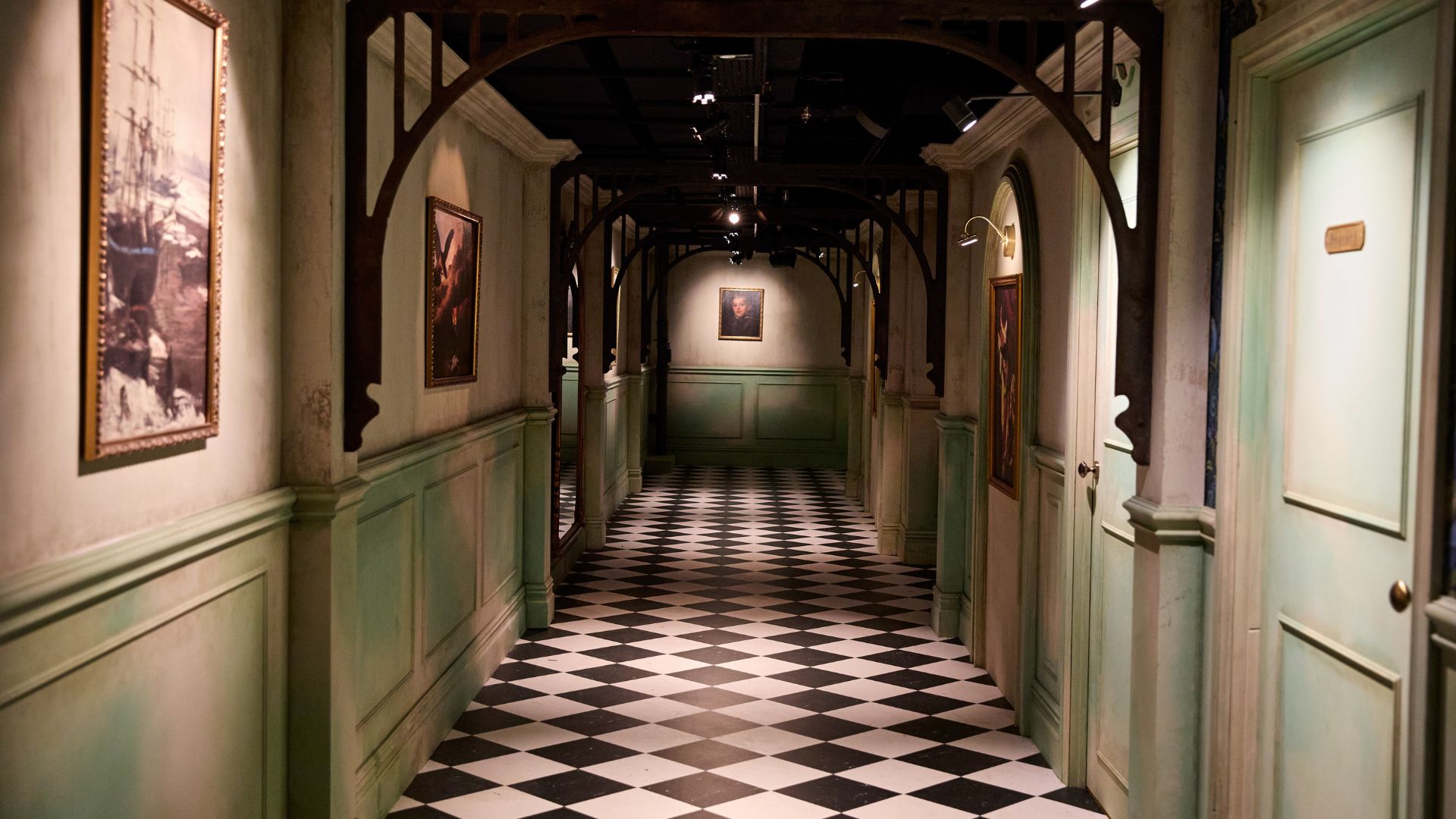 Be transported into the world of The Traitors with an interior styled as the iconic Scottish castle. Image courtesy of Mance Communications. Hallway at The Traitors: Live Experience with black and white chequered floor, antique pictures and green doorways.