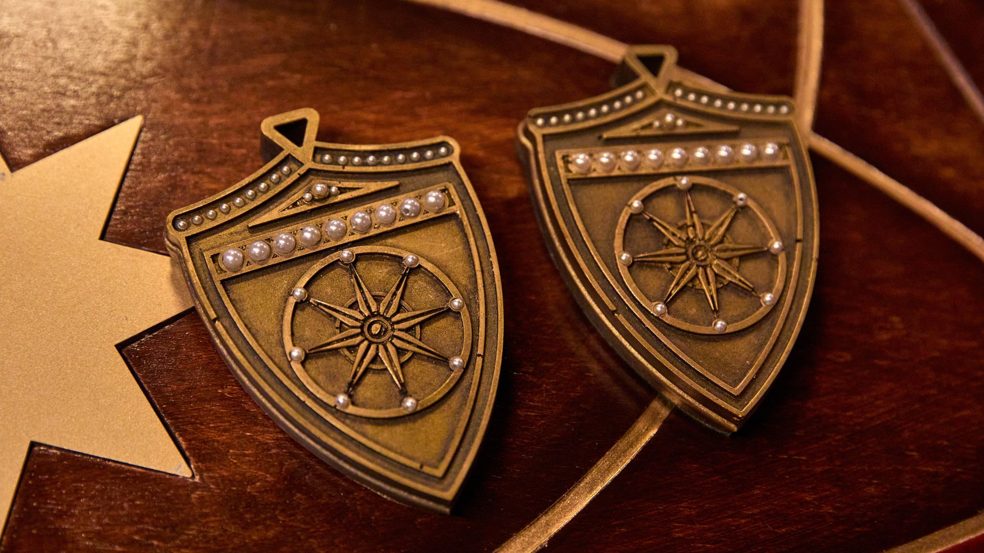 Take part in missions to win gold for the table and try to bag yourself a shield. Image courtesy of Mance Communications. Two gold shields places next to each other on wooden table.
