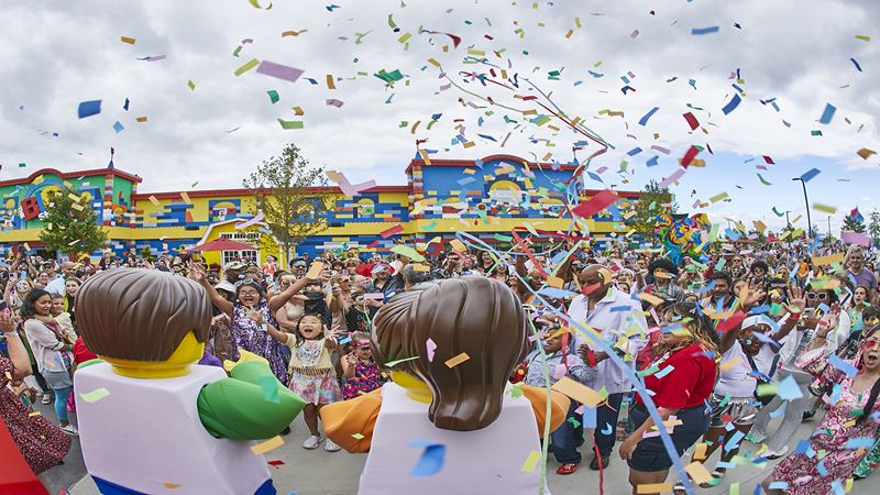 Two big Lego figures with a big Lego building in the background and a group of children in front facing the figures and confetti in the air.