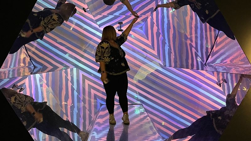 a woman stood in a mirrored and coloured kaleidoscope with her hands up 