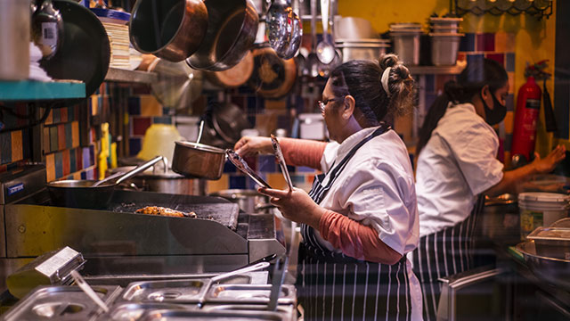 Two chefs dressed in chefs' whites and blue striped aprons work in a kitchen, one wearing a face mask, the other holding a pot in one hand and tongs in another.