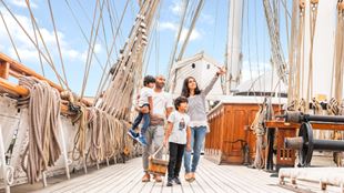 Standing on the deck of the ship Cutty Shark, a women points out to the sky, showing something to the two children and man accompanying her.