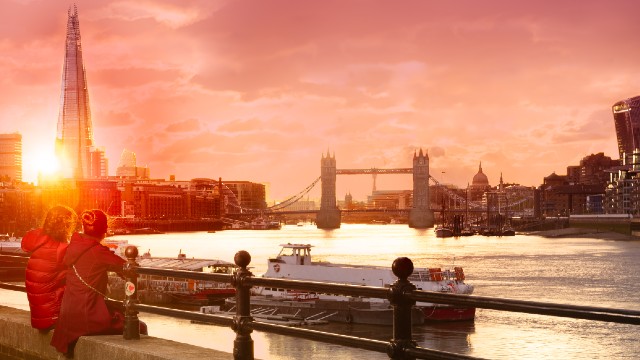 Two people sit on the banks of the river Thames to watch the sun set over the London skyline including The Shard and Tower Bridge. 