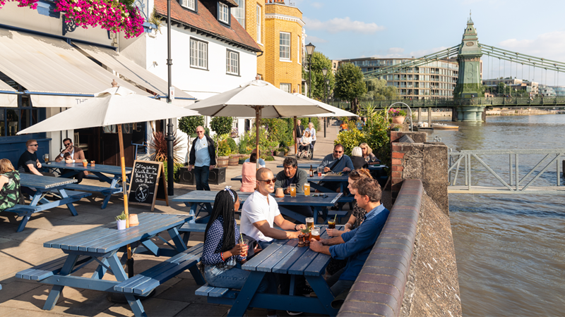 Enjoy cosy riverside pubs in Hammersmith. Credit: Antoine Buchet. Image courtesy of Visit London. Group of people enjoying drinks at a riverside pub in Hammersmith