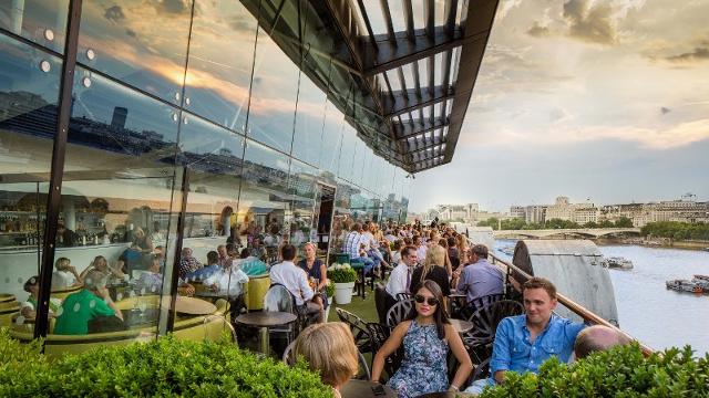 Terrace at OXO. Image courtesy of OXO Tower Restaurant, Bar and Brasserie. An outdoor rooftop overlooking the London skyline, with people eating and drinking in the hazy sunshine.