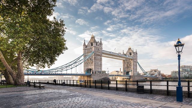 Tower Bridge in London, with a cloudy blue sky in the background and green trees in the foreground