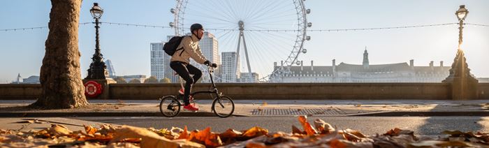 A man cycling on a bridge with the London Eye in the background and autumn leaves in the foreground.