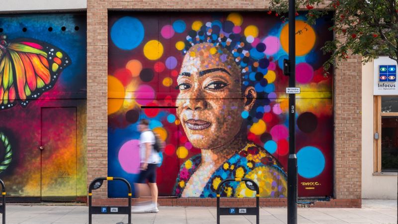 See incredible street art during the London Mural Festival. Credit: Bonzai, Otto Schade, Jimmy C. Image courtesy of Global Street Art. Colourful street art mural featuring a portrait of a black woman with bright dots of colour around her face, including blue, orange and pink splashes of colour, on brick London wall.