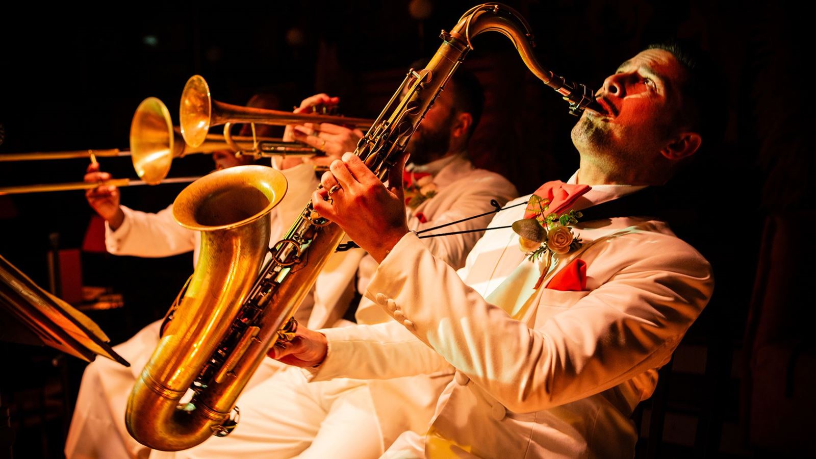 A trio of brass band players with a saxophone and trumpet in white suit jackets.