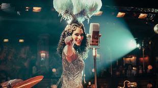 A woman dressed in a silver sparkly outfit stands next to a microphone smiling and pointing to the audience.