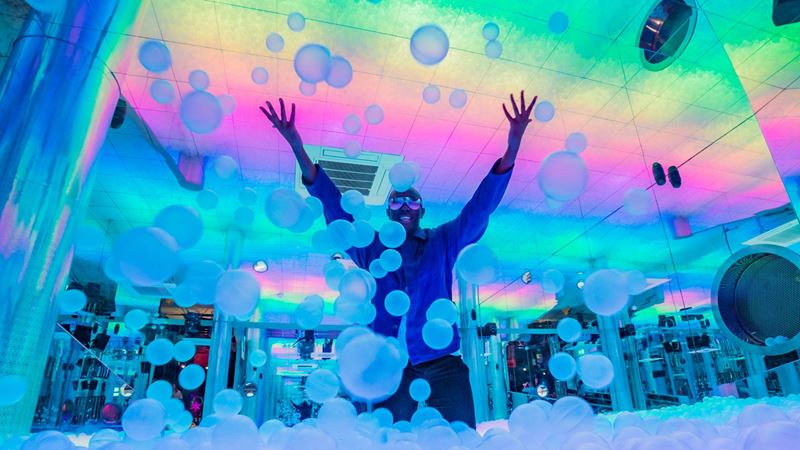 Play in Ballie Ballerson's glowing ball pit. Credit: Ballie Ballerson. Image courtesy of Ballie Ballerson. a man stood in the middle of an indoor ball pit throwing balls in the air, with bright colourful lights lighting up the room