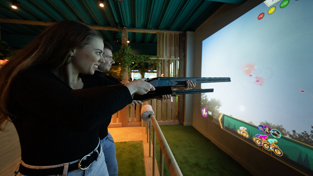 Virtual clay shooting at Clays. Image courtesy of Clays. A woman holding up a fake long barrel gun, playing a fun virtual clay pigeon shooting game.